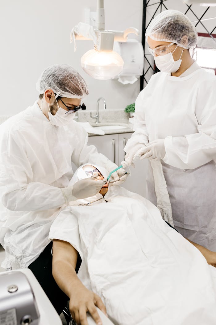 From above of anonymous orthodontist and assistant in white uniforms treating teeth of man using medical instruments in clinic